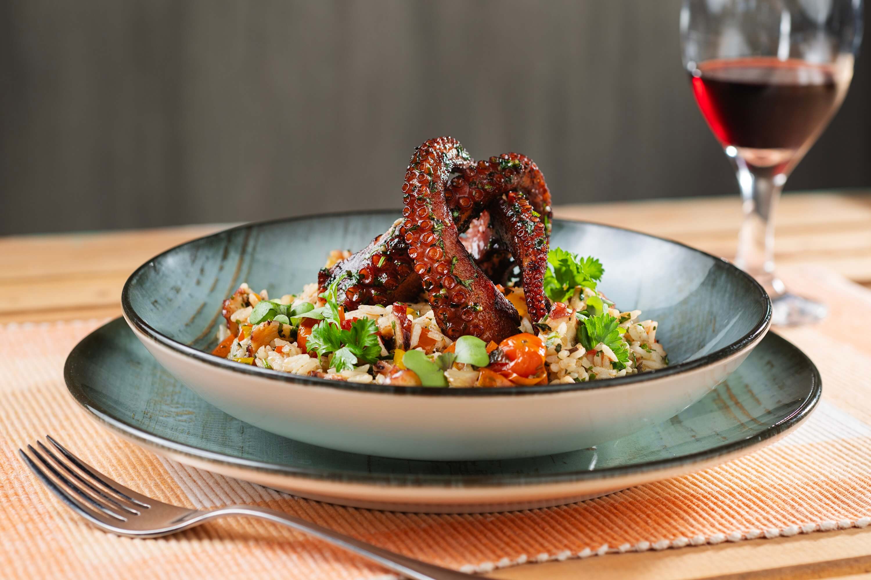 Arroz com polvo temperado com grão e legumes sobre a mesa do Restaurante Basílico, ao lado de taça de vinho tinto.